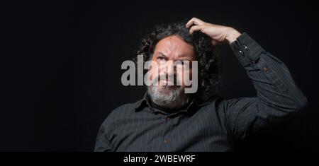 Portrait of latin man with white beard and black curly hair with doubtful expression, scratching head with hand, wearing black shirt against black bac Stock Photo
