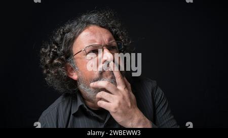 Portrait of latin man with white beard and black curly hair with expression of indecision, looking to the right, wearing black shirt against black bac Stock Photo