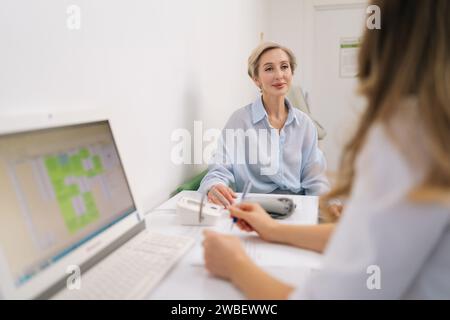 Rear view of unrecognizable female doctor in white uniform to middle-aged woman patient, discuss health concerns writes complaints, fill medical form. Stock Photo