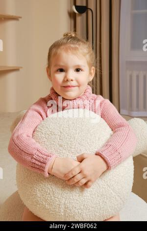 In a cozy room, little girl in pink sweater embraces a large white stuffed decorative pillow Stock Photo