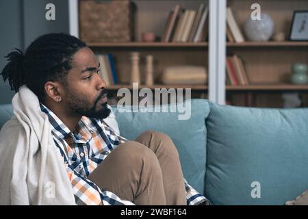 Young depressed African American man sitting on sofa with crossed arms. Racism victim for skin color black male suffering from depression after being Stock Photo