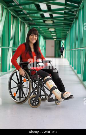 A young woman with a leg in plaster sits in a wheelchair and smiles, hospital Stock Photo