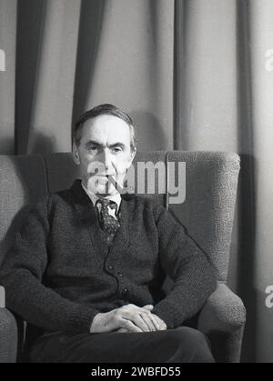 1960s, historical, a middle-aged man wearing a  shirt & tie and wool cardigan sitting in a lounge chair, raylon covered fabric of the era, a pipe in his mouth, looking at the camera, England, UK. Stock Photo