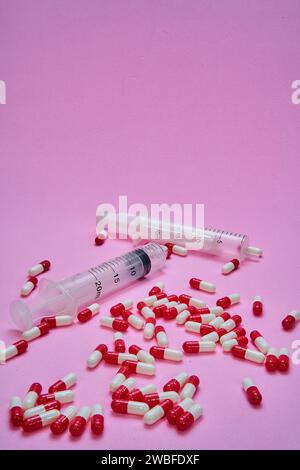 Two syringes and a pile of red and white pills and capsules on a pink table. Stock Photo