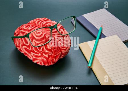 Representation of a brain with glasses and a notebook and pencil on a dark-colored table. Stock Photo