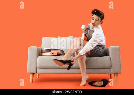 Young pin-up businesswoman with coffee and folders taking off heels on sofa against orange background Stock Photo