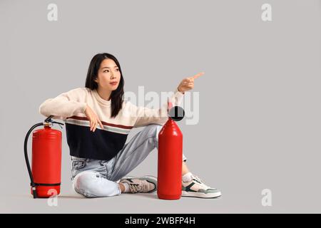 Young Asian woman with fire extinguishers pointing at something while sitting against grey background Stock Photo