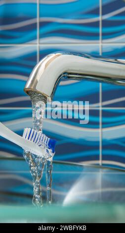 Close-up view of a white and blue toothbrush being washed under a stream of water coming out of a chrome metal faucet against a blue ceramic wall back Stock Photo