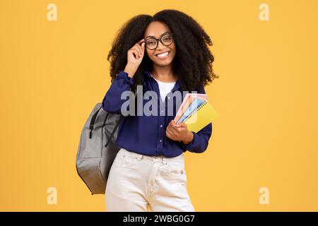 Happy black student with curly hair, backpack and copybooks, adjusting eyeglasses, poses on yellow background Stock Photo