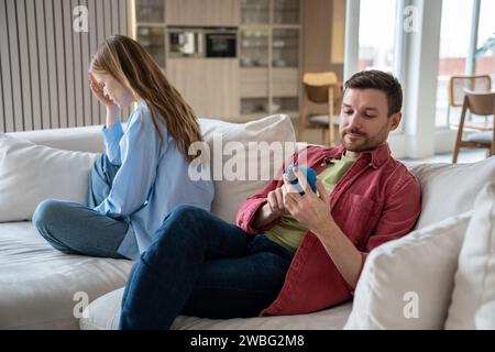Husband ignoring wife emotions looking at smartphone screen sitting on couch at home.  Stock Photo