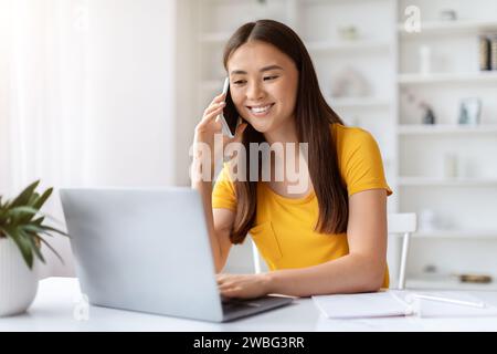 Young asian woman smiling while talking on cellphone and working on laptop Stock Photo