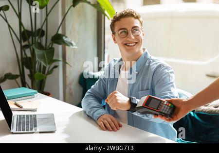 Happy young european guy using smartwatch for payment machine, sit on desk with laptop Stock Photo