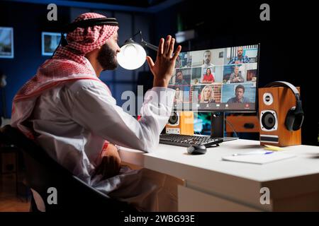 Arab businessman sitting at his office desk having a video conference call with group of coworkers. Young Muslim guy using his desktop pc to conduct an online meeting with different people. Stock Photo