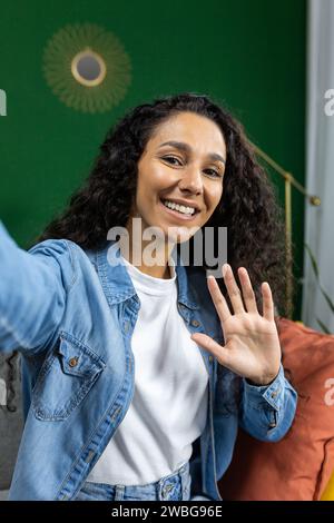 Vertical shot, woman at home on sofa looking at phone camera smiling and talking with friends using smartphone app, waving greeting gesture and taking selfie photo, in living room. Stock Photo