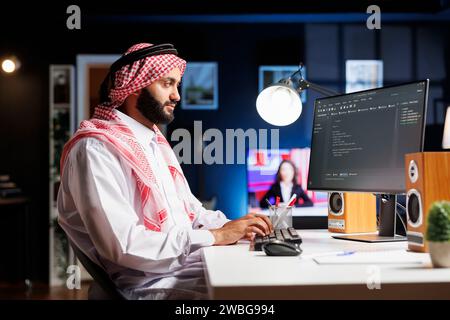 Side-view of Muslim developer writing code on desktop pc, parsing algorithm in a software agency. Using a computer monitor, a Middle Eastern male coder works on a user interface. Stock Photo