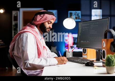 Muslim man sits at a computer and programs, exhibiting the app development process. Side-view image of a computer monitor displaying algorithms created by an Arab guy dressed traditionally. Stock Photo