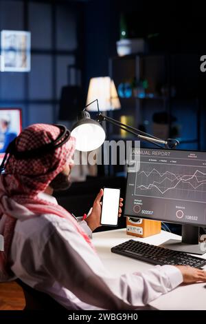 Muslim man sits at office desk, holding mobile device with blank white screen. Arab guy vertically grasping smartphone displaying isolated mockup template. Ideal for advertising or software templates. Stock Photo