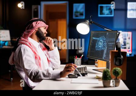 Middle Eastern professional in Arabic attire works at monitoring a secure cloud-based network with a desktop computer. He utilizes AI and machine learning for efficient and seamless online services. Stock Photo