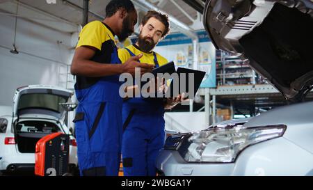 Repair shop employees work together on fixing car, using tablet and laptop. Teamworking colleagues collaborating on servicing broken vehicle, checking for defective components using electronic devices Stock Photo