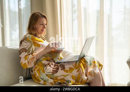 Pregnant woman, in a flowered dress, drinking tea and using a laptop sitting in the hotel hall.  Stock Photo