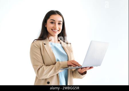 Business concept. Beautiful confident indian or arabian business woman, office worker in elegant suit, holding an open laptop, standing on isolated white background, looks at camera, smiling friendly Stock Photo