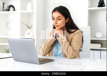 Serious concentrated young adult indian or arabian business lady, executive, company employee, lawyer, in formal wear, sitting in modern office, focused looks at laptop screen, studying information Stock Photo