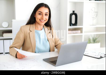 Cute attractive young adult indian or arabian business woman entrepreneur, financial expert of company, wearing suit, sitting in the office, holding financial documents, looking at camera and smiles Stock Photo
