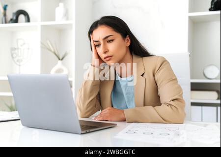 Tired overworked young adult indian or arabian business lady, company employee, lawyer, in formal wear, sitting in modern office, looks tiredly at the laptop screen, has a work deadline, need rest Stock Photo