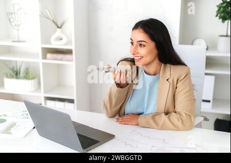 Attractive young woman of indian or arabian nationality, sit at desk holding smart phone and talk on speakerphone, share information, record messages to clients, smiling. Speakerphone concept Stock Photo