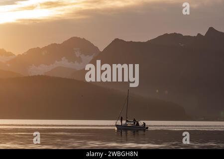 sailboat sails through calm waters beneath snow capped mounting in golden morning sunrise light Stock Photo