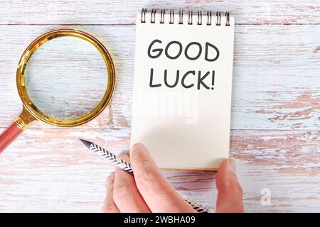 Good luck written on a blank sheet of a notebook which is touched by a man's hand with a pencil and a magnifying glass is lying next to it Stock Photo