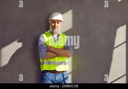 Portrait of mature bearded foreman in a helmet and construction vest on grey wall background. Stock Photo