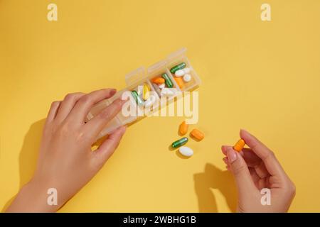 Against the yellow background, female hands is holding the plastic box containing pills of various colors and shapes by day for memory. Daily pill sch Stock Photo