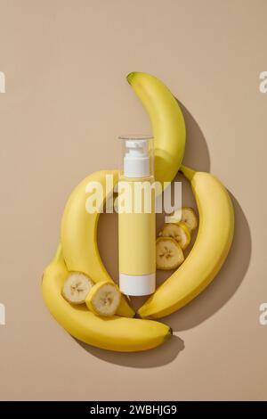 Cosmetic bottle in yellow color is decorated with several fresh bananas. Beige background. Banana (Musaceae) is one of the most widely consumed fruits Stock Photo