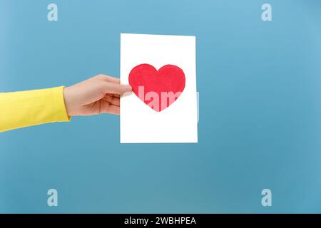 Close up of female hand holding card with heart symbol, posing isolated over plain blue color background wall in studio with copy space for advertisem Stock Photo
