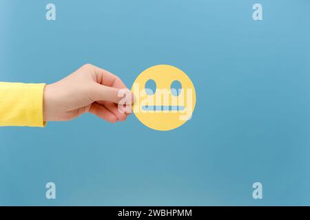 Close up of female hand holding yellow emoticon with boring face, posing isolated over plain blue color background wall in studio with copy space for Stock Photo