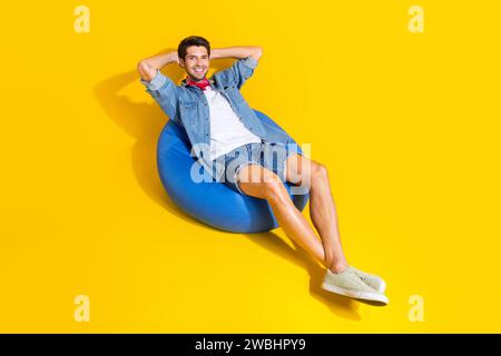 Photo of relaxed satisfied guy wear jeans jacket shorts lie on pouf hold arms behind head isolated on bright yellow color background Stock Photo