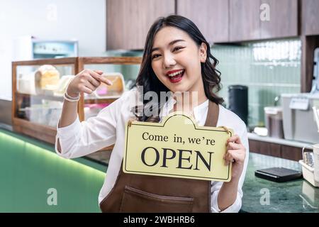 Happy beautiful Asian female waitress or younger shopkeeper in an apron holding open sign while standing at cafe or in restaurant Stock Photo