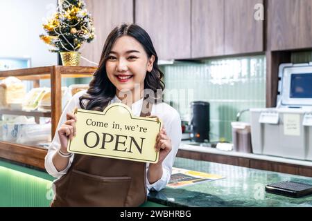 Happy beautiful Asian female waitress or younger shopkeeper in an apron holding open sign while standing at cafe or in restaurant Stock Photo