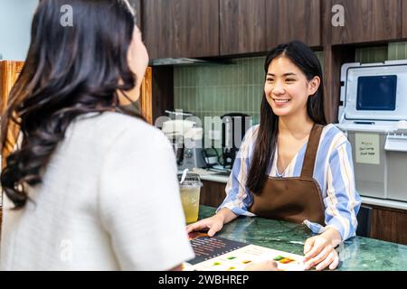 Smiling young asian waitress in a cafe with customer for service, payment or order on counter at coffee shop. Stock Photo
