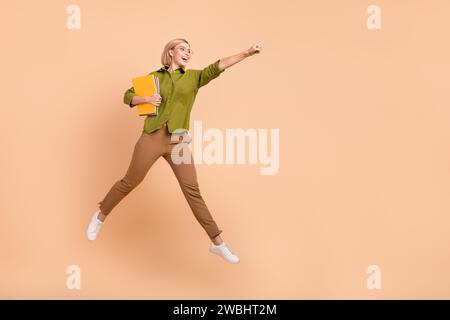 Full size photo of nice girl wear khaki shirt flying hold copybooks clenching fist look empty space isolated on pastel color background Stock Photo
