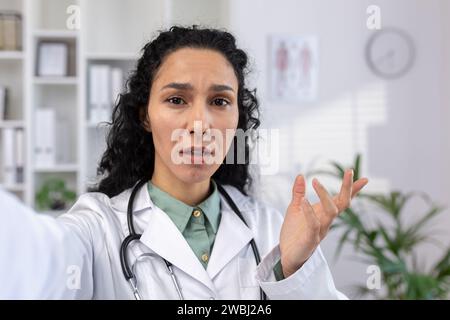 Female doctor is serious, looking worriedly into smartphone camera talking to patient remotely, reporting disappointing results online, Hispanic woman in white medical coat using app on phone video call. Stock Photo