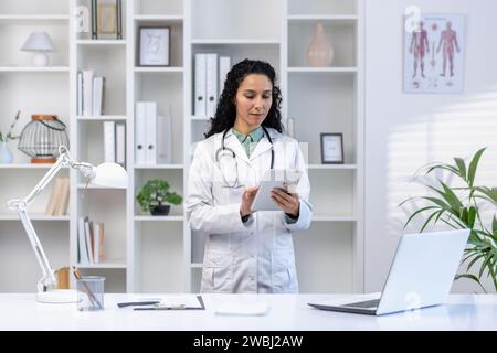 Senior mature female doctor serious and confident thoughtfully using tablet computer, Latin American woman in medical gown standing inside clinic office near desk, using app. Stock Photo
