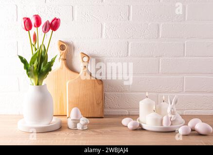 Easter background, front view on table decorated with flowers in a vase, traditional eggs and ceramic rabbits, space for a text. white brick wall. Stock Photo