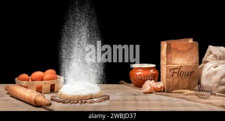 Cooking bread. The dough lies on a wooden kitchen table and sprinkled with flour on top. On the kitchen board are eggs in a basket, a bag of flour, a Stock Photo