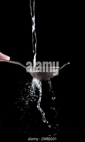 A stream of water pours through a small steel sieve in the hands of a man on a black background. Splashes of water fly out through a sieve. Stock Photo