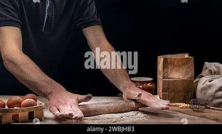 Man cooks homemade bread in the kitchen. Baker rolls dough with a rolling pin on a wooden table sprinkled with flour Stock Photo