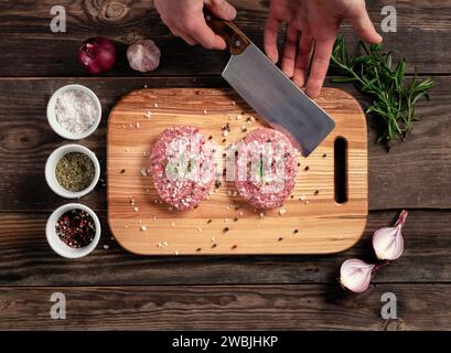 Fresh raw organic cutlets for homemade burgers. The cook picks up a meat cutlet with a knife. On wooden worktop placed plates with seasonings, garlic, Stock Photo