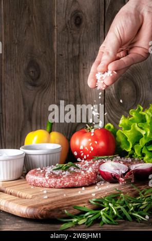 Raw organic meat cutlets. Cook sprinkles salt on meat cutlets on a wooden cutting board. Preparing meat for a delicious homemade hamburger. Stock Photo