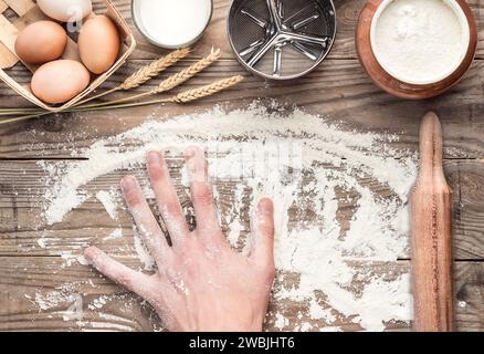 Top view of a wooden kitchen worktop. Flour is scattered on the countertop. On the table are a basket of eggs, a glass of milk, a sieve mug, a rolling Stock Photo